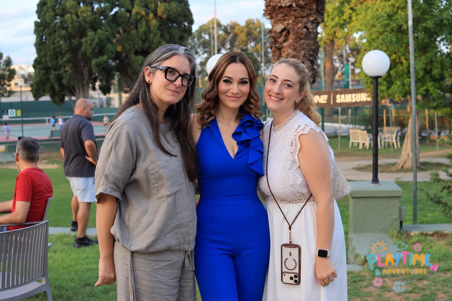 Three friends smiling by the tennis courts at the Summer Party